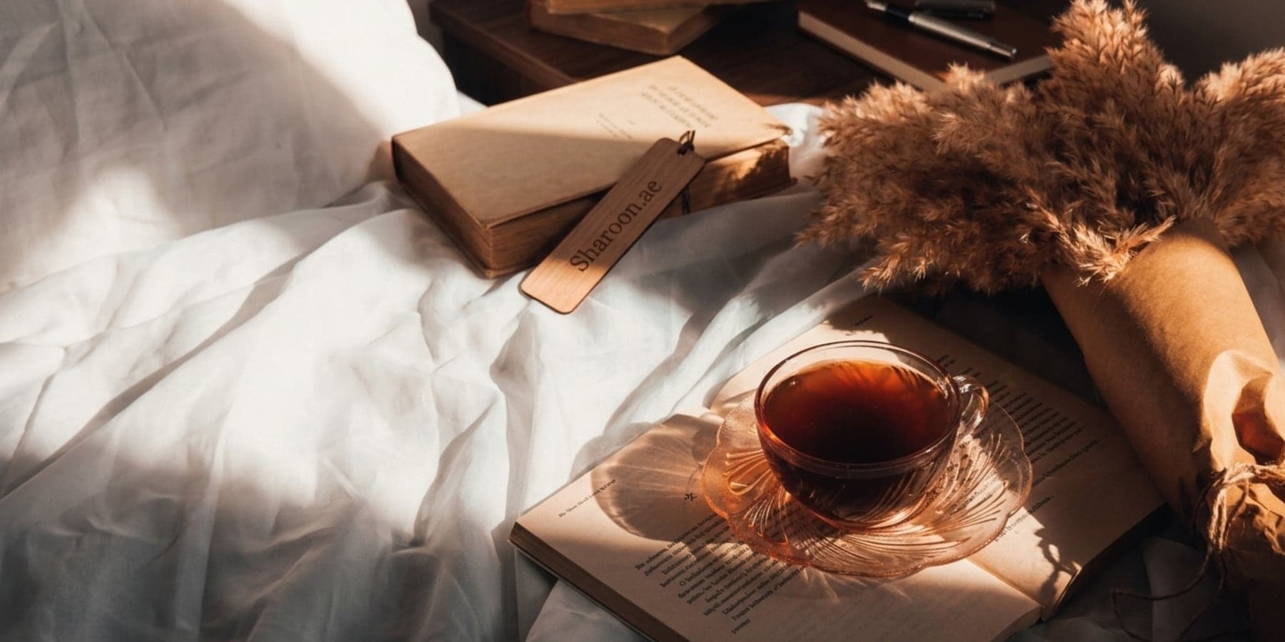 A Sharoon.ae bookmark resting on linen beside tea and an open book